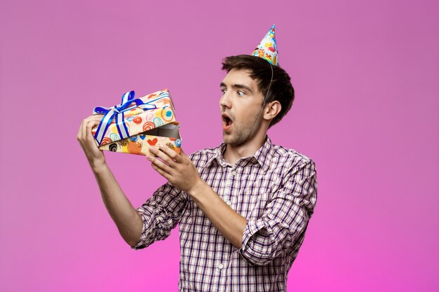 Sorprendido joven guapo abriendo regalo de cumpleaños sobre pared púrpura.