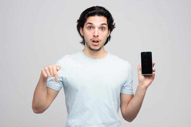 Sorprendido joven guapo mostrando teléfono móvil mirando a la cámara apuntando hacia abajo aislado sobre fondo blanco.