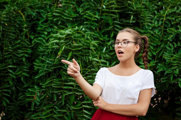 Sorprendido joven hermosa estudiante en gafas apuntando con el dedo