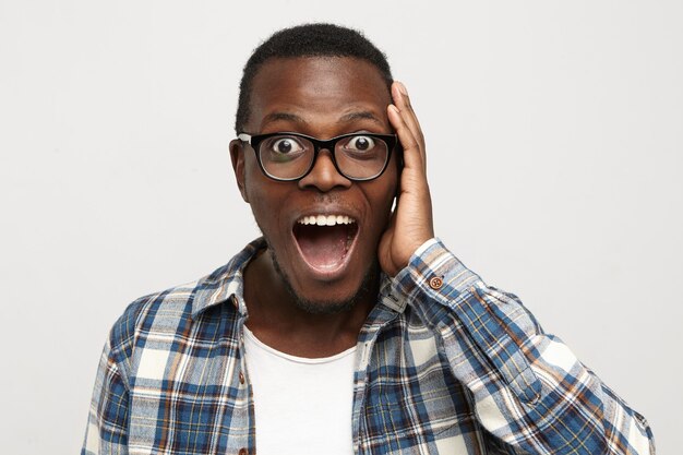 Sorprendido joven inconformista afroamericano con gafas y camisa a cuadros sobre camiseta blanca