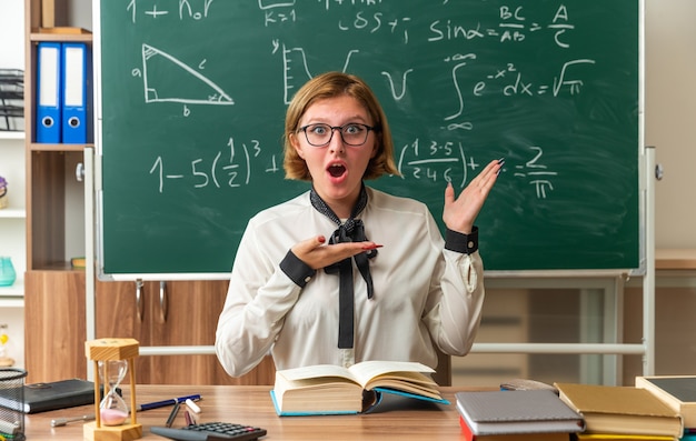 Sorprendido joven maestra se sienta a la mesa con útiles escolares puntos al lado en el aula