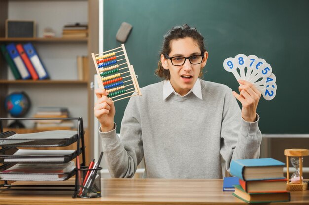 sorprendido joven maestro usando anteojos sosteniendo abacus con número divertido sentado en el escritorio con herramientas escolares en el aula