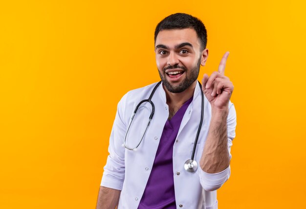 Sorprendido joven médico con estetoscopio bata médica apunta al lado con el dedo en la pared amarilla aislada