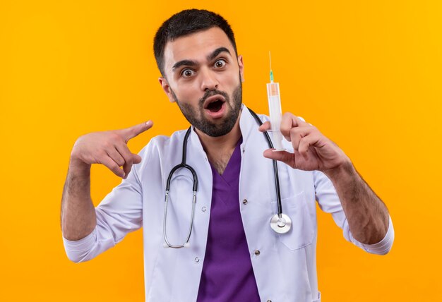 Sorprendido joven médico con estetoscopio bata médica apunta a la jeringa en la mano en la pared amarilla aislada