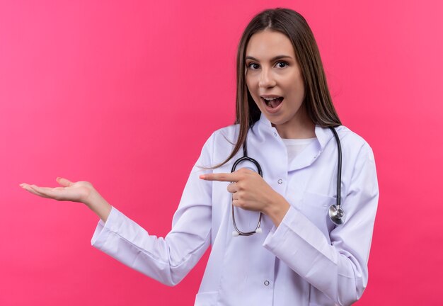 Sorprendido joven médico con estetoscopio bata médica puntos a mano sobre fondo rosa aislado