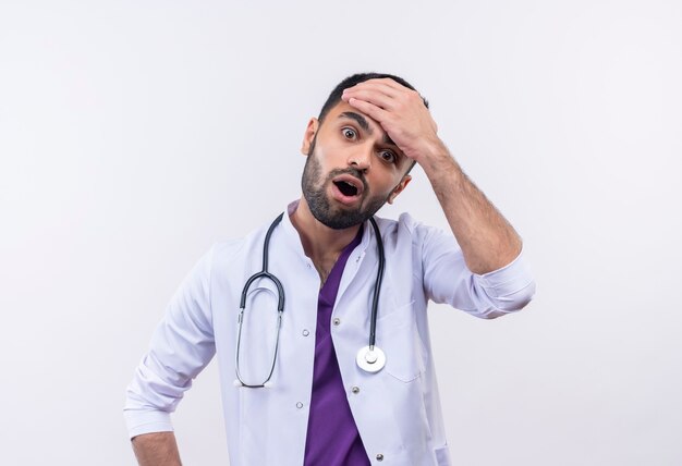 Sorprendido joven médico con estetoscopio bata médica puso su mano en la frente en blanco aislado