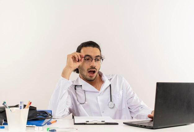 Sorprendido joven médico con gafas médicas vistiendo bata médica con estetoscopio