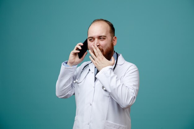 Sorprendido joven médico masculino con abrigo médico y estetoscopio alrededor del cuello hablando por teléfono mirando a un lado mientras mantiene la mano en la boca aislada en el fondo azul