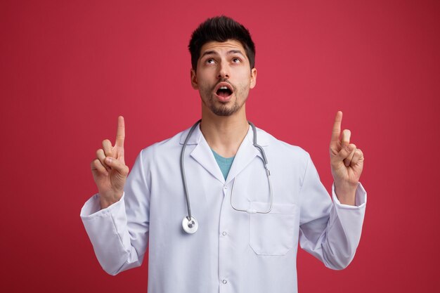 Sorprendido joven médico masculino con uniforme médico y estetoscopio alrededor del cuello mirando hacia arriba señalando con el dedo aislado en el fondo rojo