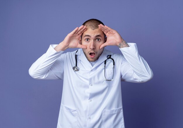 Sorprendido joven médico vistiendo una bata médica y un estetoscopio alrededor de su cuello haciendo grandes ojos aislados en la pared púrpura