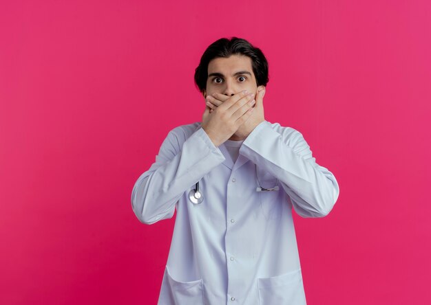 Sorprendido joven médico vistiendo una bata médica y un estetoscopio manteniendo las manos en la boca aislada en la pared rosa con espacio de copia