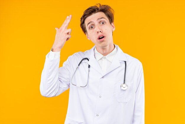 Sorprendido joven médico vistiendo bata médica con estetoscopio mostrando gesto de pistola