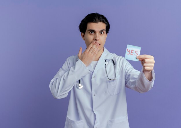 Sorprendido joven médico vistiendo bata médica y estetoscopio mostrando sí nota manteniendo la mano en la boca aislada en la pared púrpura con espacio de copia