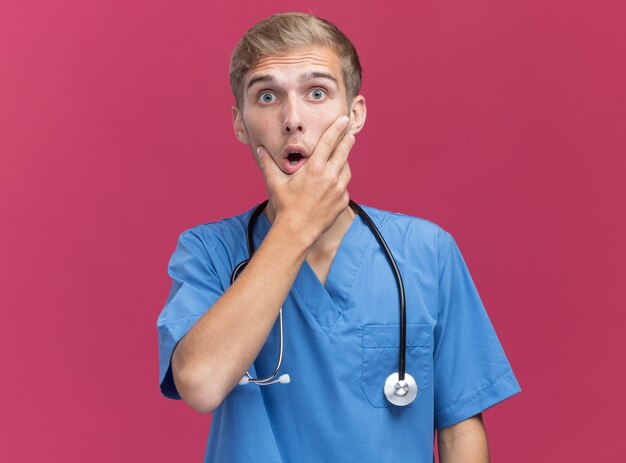 Sorprendido joven médico vistiendo uniforme médico con estetoscopio agarró la barbilla aislada en la pared rosa