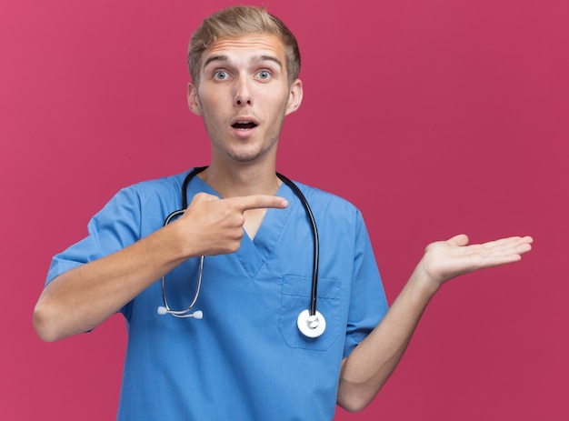 Sorprendido joven médico vistiendo uniforme médico con estetoscopio fingiendo sostener y apunta a algo aislado en la pared rosa con espacio de copia