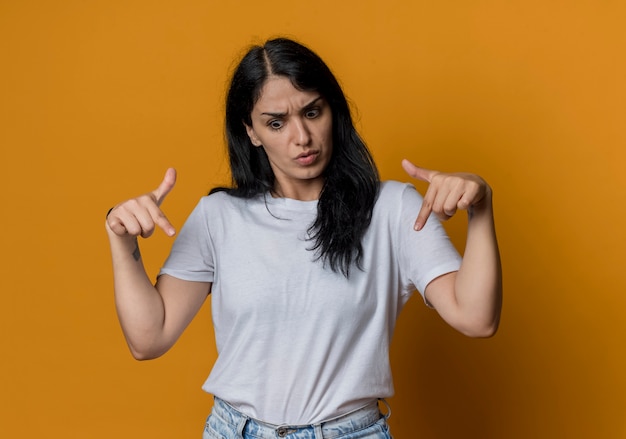 Sorprendido joven morena caucásica apunta hacia abajo aislado en la pared naranja