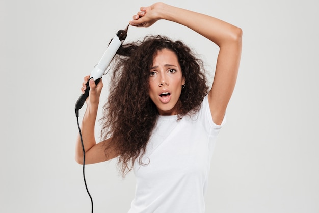 Sorprendido joven mujer morena enderezar su cabello y mirando la cámara aislada sobre blanco