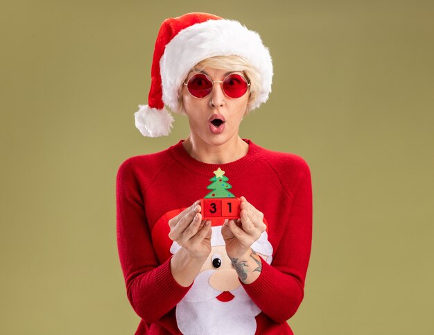 Foto gratuita sorprendido joven mujer rubia con sombrero de navidad y santa claus suéter de navidad con gafas sosteniendo el juguete del árbol de navidad con fecha mirando aislado en la pared verde oliva con espacio de copia