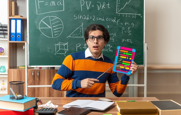 Sorprendido joven profesor de geometría caucásico con gafas sentado en un escritorio con útiles escolares en el aula mostrando el ábaco apuntando con el puntero mirando al frente