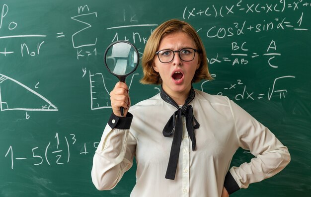 Sorprendido joven profesora con gafas de pie delante de la pizarra sosteniendo lupa poniendo la mano en la cadera en el aula