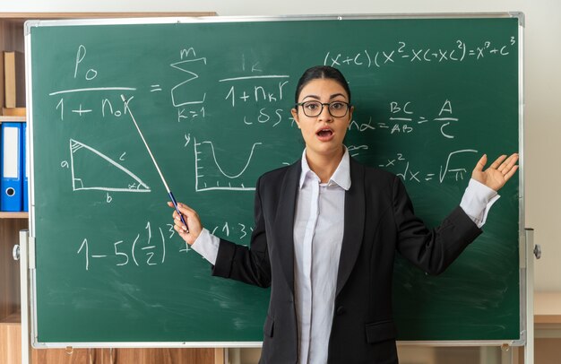 Sorprendido joven profesora con gafas de pie delante de la pizarra sosteniendo el palillo del puntero extendiendo las manos en el aula