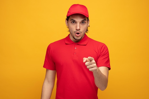 Sorprendido joven repartidor con uniforme y gorra mirando y apuntando a la cámara aislada de fondo amarillo