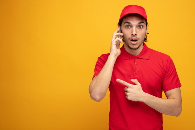 Sorprendido joven repartidor con uniforme y gorra mirando a la cámara apuntando al lado aislado en fondo amarillo con espacio para copiar