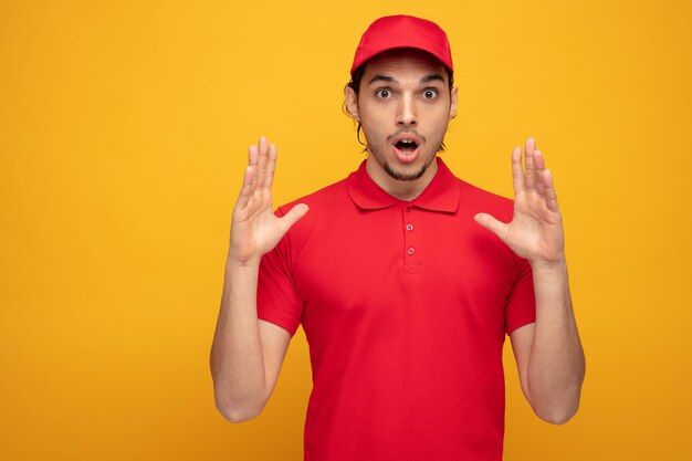 Sorprendido joven repartidor con uniforme y gorra mirando a la cámara mientras mantiene las manos en el aire aisladas en el fondo amarillo