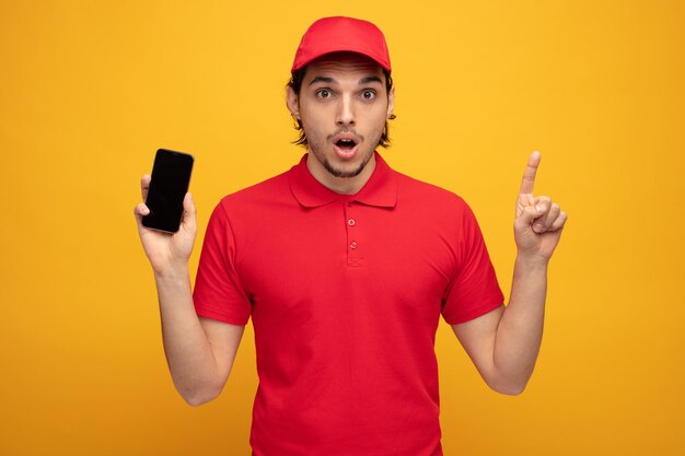 sorprendido joven repartidor con uniforme y gorra que muestra el teléfono móvil mirando a la cámara apuntando hacia arriba aislado en fondo amarillo