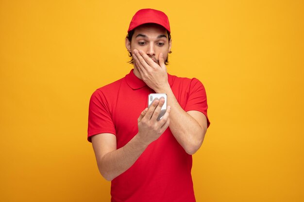 sorprendido joven repartidor con uniforme y gorra sosteniendo y mirando el teléfono móvil manteniendo la mano en la boca aislada sobre fondo amarillo