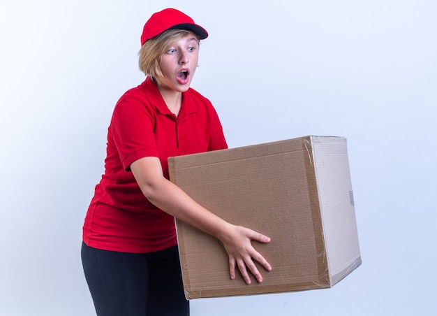 Sorprendido joven repartidora vistiendo uniforme y gorra con caja aislada en la pared blanca