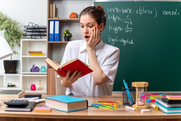 Sorprendido joven rubia profesora de matemáticas sentada en el escritorio con herramientas escolares leyendo el libro manteniendo la mano en la cara en el aula