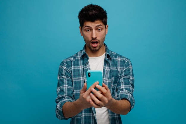 Sorprendido joven sosteniendo y mirando el teléfono móvil aislado sobre fondo azul.