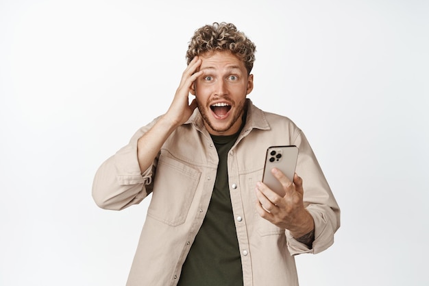 Sorprendido joven sosteniendo un teléfono móvil mirando con incredulidad a la cámara asombrado por algo en la aplicación de teléfono inteligente de pie contra el fondo blanco