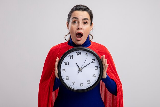 Sorprendido joven superhéroe con reloj de pared aislado sobre fondo blanco.