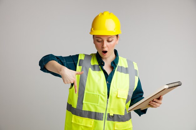 Sorprendido joven trabajador de la construcción con casco de seguridad y chaleco de seguridad sosteniendo el bloc de notas mirando y apuntando hacia abajo