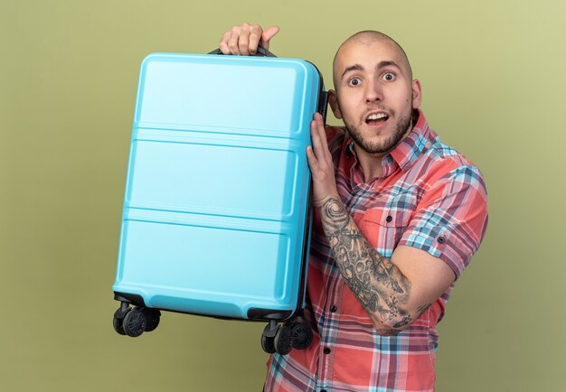 Sorprendido joven viajero hombre sujetando la maleta aislado en la pared verde oliva con espacio de copia