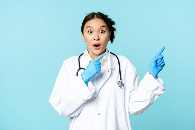 Sorprendido médico asiático enfermera en uniforme apuntando a la derecha y jadeando sorprendido gran noticia de pie sobre bl ...