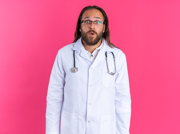 Sorprendido médico varón adulto vistiendo bata médica y estetoscopio con gafas mirando a cámara aislada en pared rosa