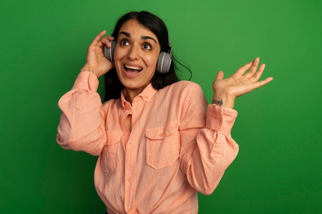 Sorprendido mirando al lado hermosa joven vestida con camiseta rosa con auriculares extendiendo la mano aislada en la pared verde