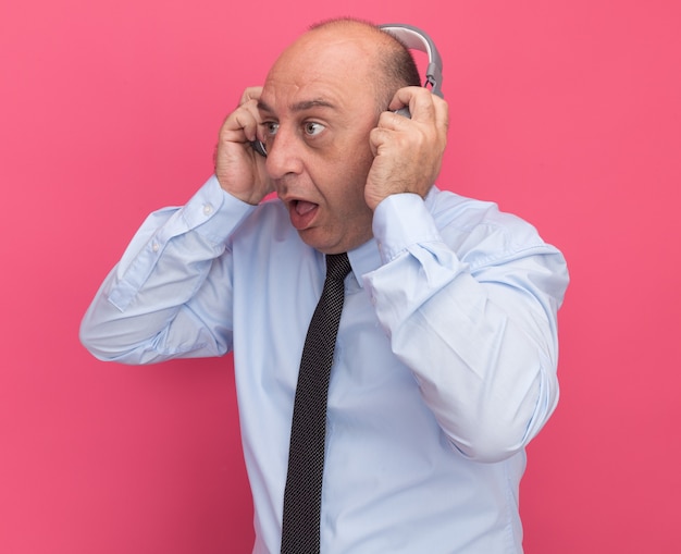 Sorprendido mirando al lado del hombre de mediana edad con camiseta blanca con corbata y auriculares aislados en la pared rosa