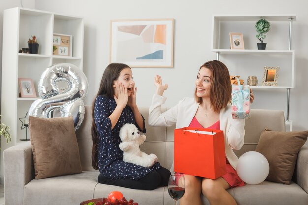 Sorprendido mirando el uno al otro niña y madre con presente y oso de peluche en el día de la mujer feliz sentado en el sofá en la sala de estar