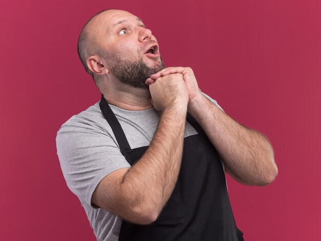 Sorprendido mirando hacia arriba barbero masculino de mediana edad eslavo en uniforme tomados de la mano juntos aislado en la pared rosa