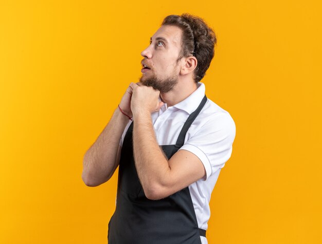 Sorprendido mirando hacia arriba el joven peluquero vistiendo uniforme poniendo las manos debajo de la barbilla aislado sobre fondo amarillo