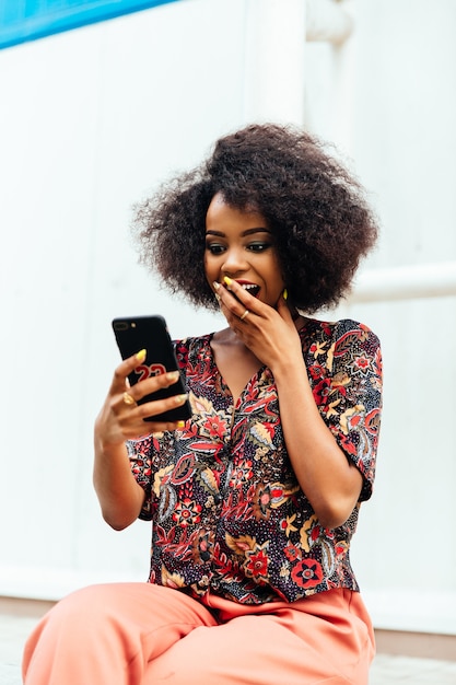 Sorprendido mujer africana, cubriendo su boca con la mano mientras mira la pantalla del teléfono inteligente