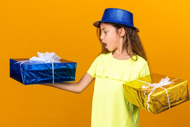 Sorprendido niña caucásica con gorro de fiesta azul sosteniendo y mirando cajas de regalo aisladas en la pared naranja con espacio de copia