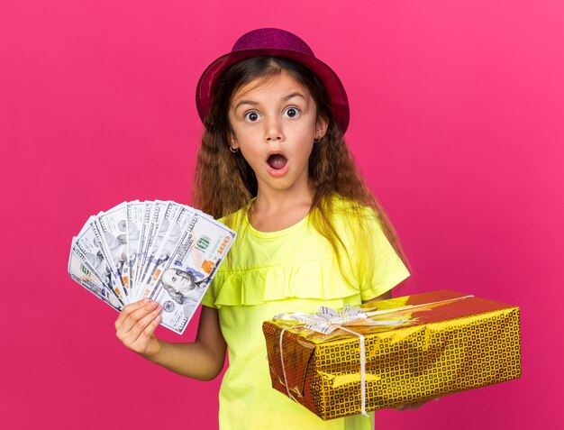 Sorprendido niña caucásica con gorro de fiesta púrpura con caja de regalo y dinero aislado en la pared rosa con espacio de copia