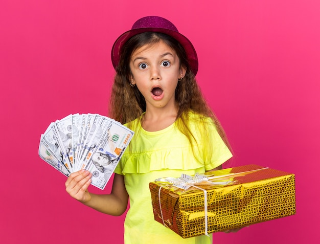 Sorprendido niña caucásica con gorro de fiesta púrpura con caja de regalo y dinero aislado en la pared rosa con espacio de copia