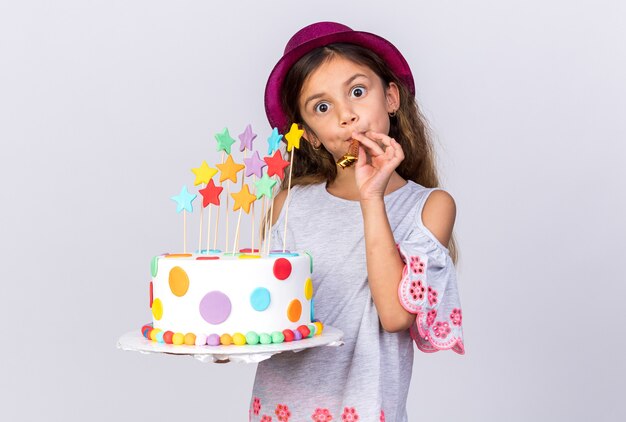 Sorprendido niña caucásica con gorro de fiesta púrpura sosteniendo pastel de cumpleaños y soplando silbato de fiesta aislado en la pared blanca con espacio de copia