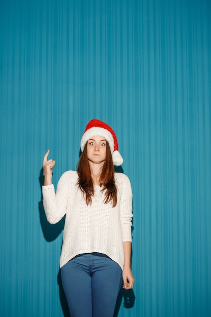 Sorprendido niña de Navidad con un gorro de Papá Noel apuntando hacia arriba sobre el fondo azul del estudio
